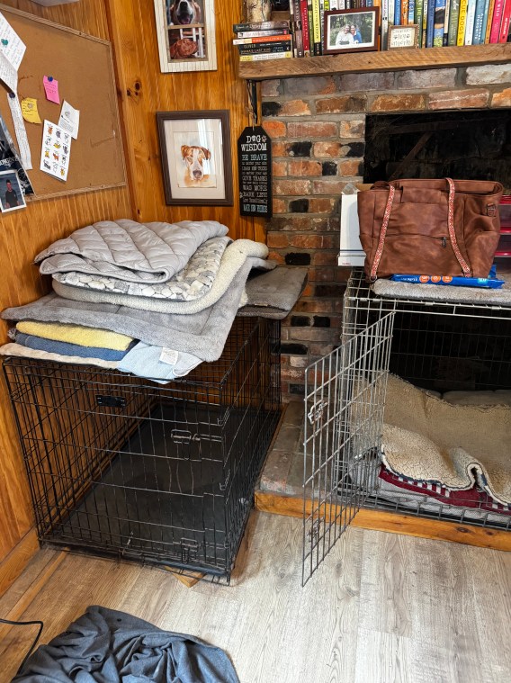 A cozy living area featuring two dog crates stacked with blankets, a leather bag on top of one crate, and a brick fireplace in the background.